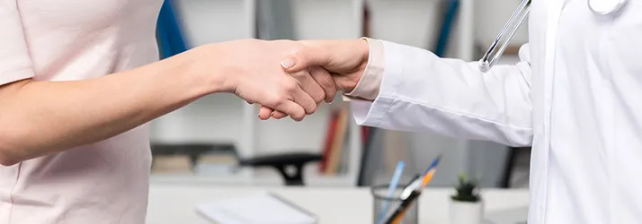 Close-up of two people shaking hands across a desk in a clinical or office setting.